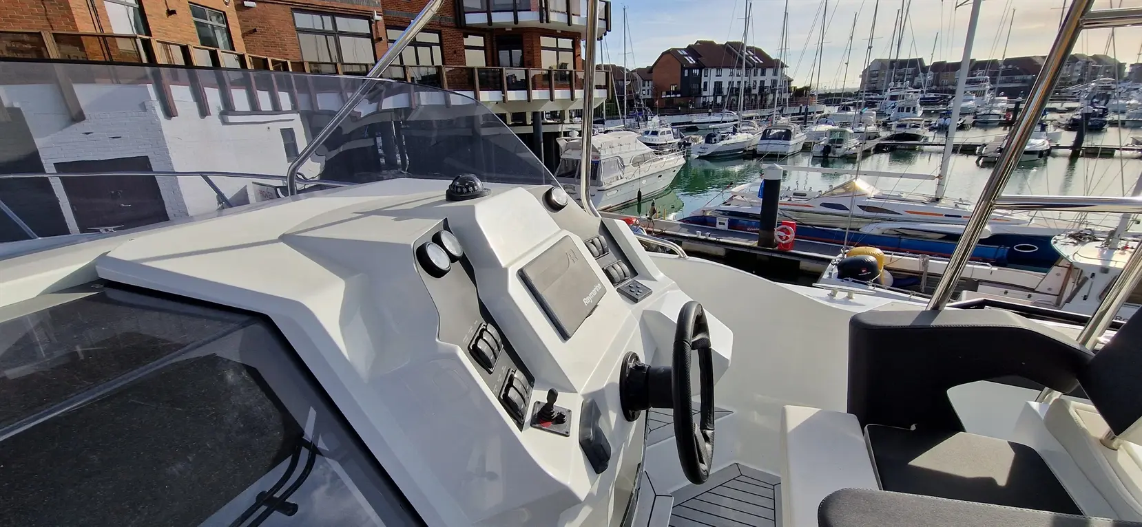 Control panel and steering wheel on a boat, overlooking a marina filled with various boats and waterfront buildings.